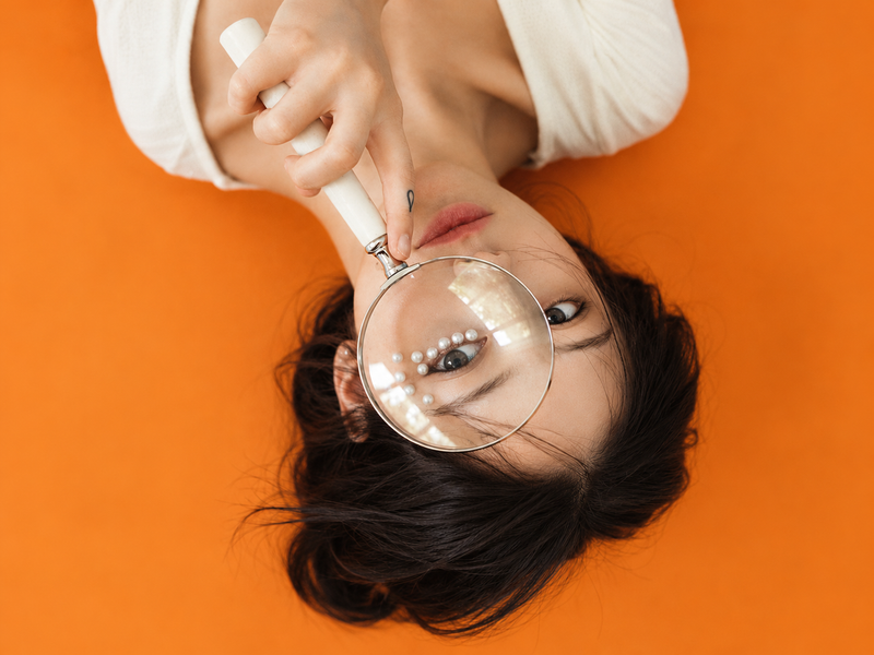 Jeune femme allongée sur fond orange, une loupe devant l'œil