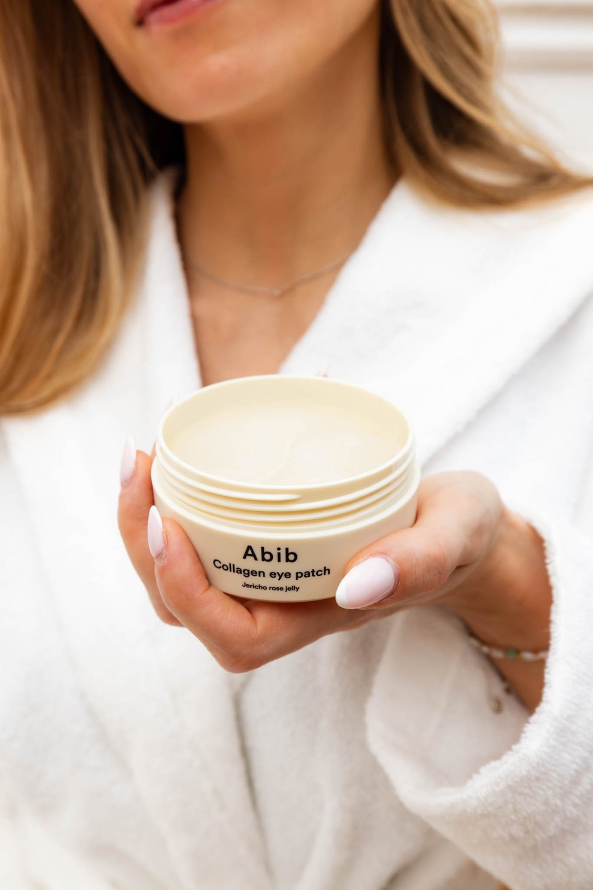 Patchs yeux Collagen Jericho Rose Jelly Abib, pot ouvert tenu en main devant un peignoir blanc