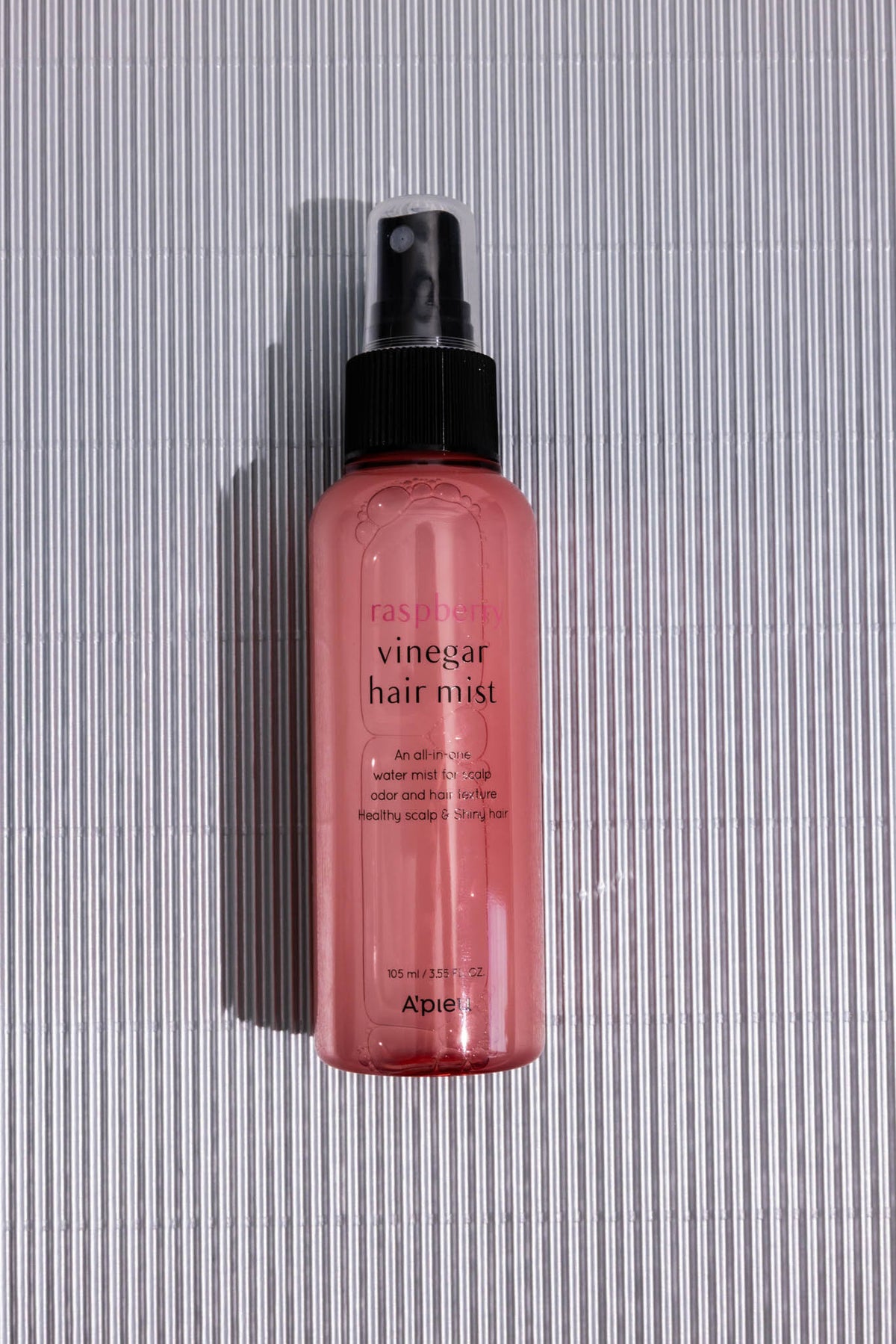 Brume cheveux Raspberry Vinegar A’PIEU, flacon spray rose posé à plat sur fond rayé gris