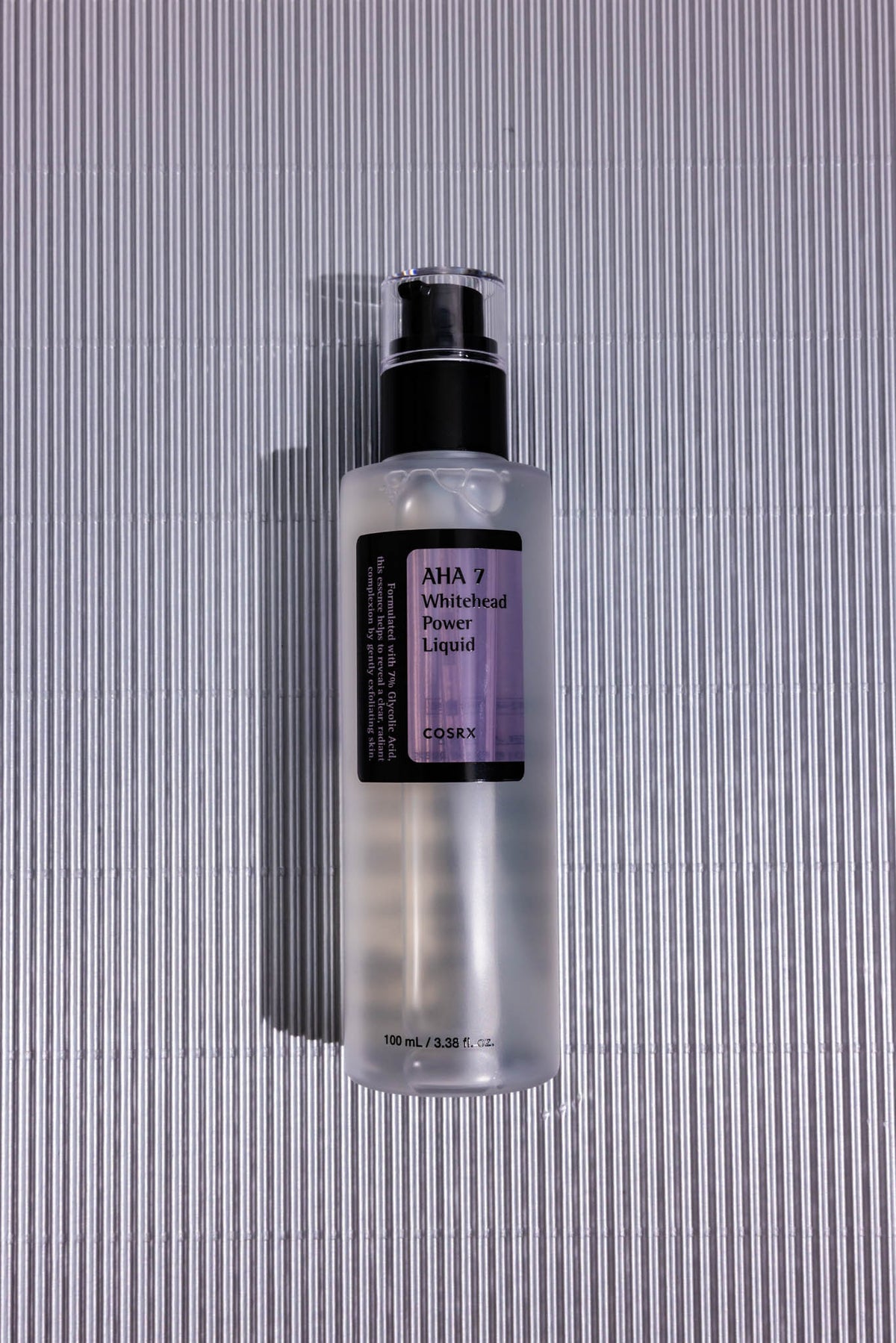 Exfoliant AHA 7 Whitehead Power Liquid COSRX, flacon pompe posé sur fond strié gris, vue de dessus
