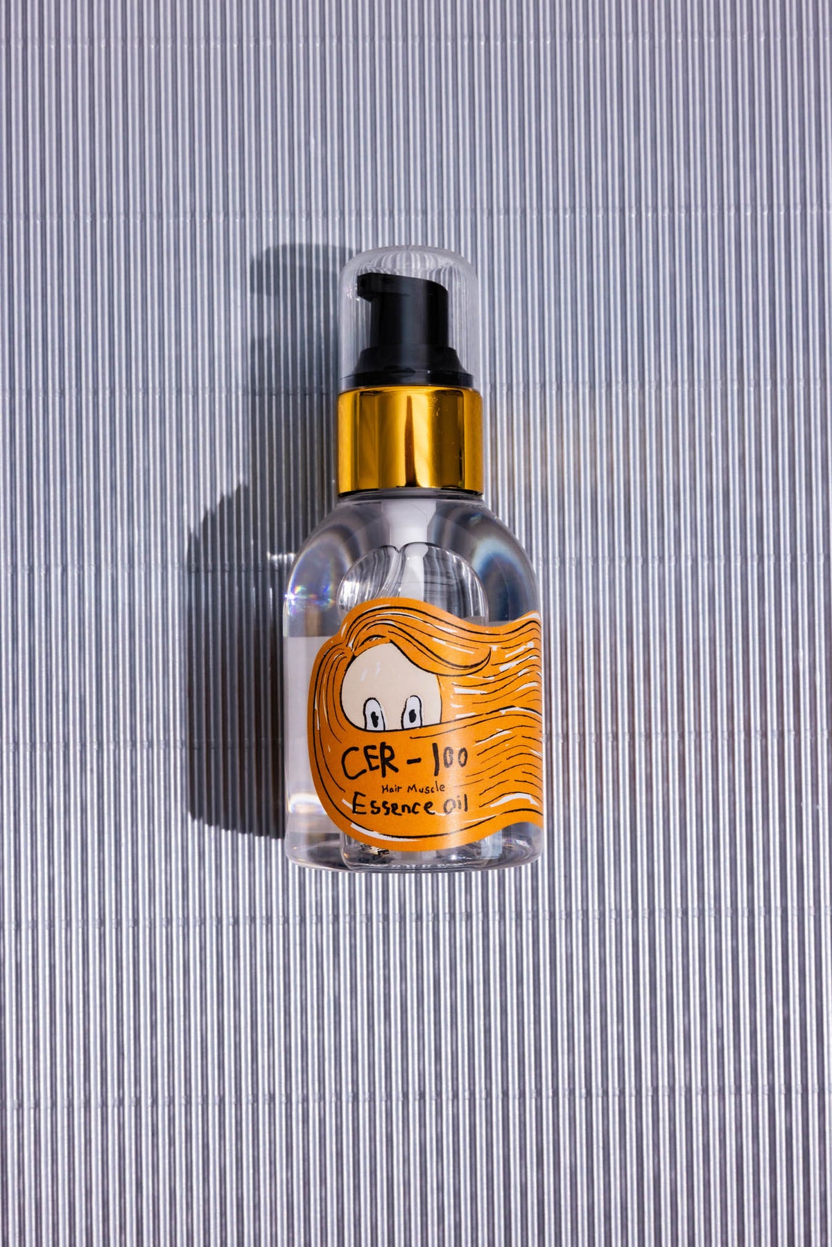 Huile cheveux Cer-100 Hair Muscle Essence Elizavecca, flacon pompe posé à plat sur fond rayé gris