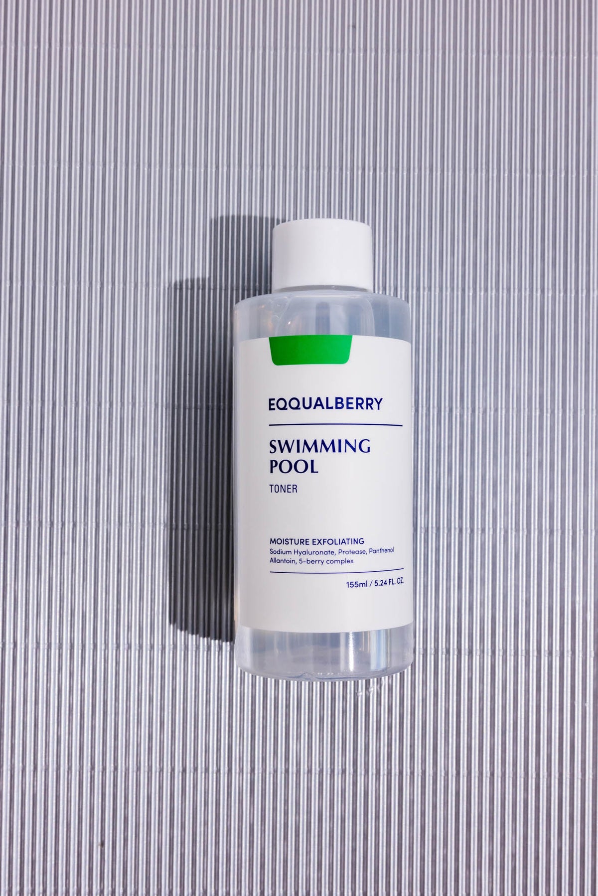 Lotion tonique Swimming Pool Daily Eqqualberry, flacon posé sur fond strié gris, vue de dessus