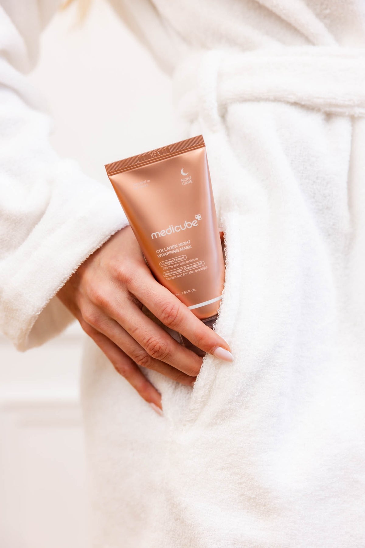 Masque de nuit Collagen Night Wrapping Mask Medicube, tube tenu en main contre un peignoir blanc