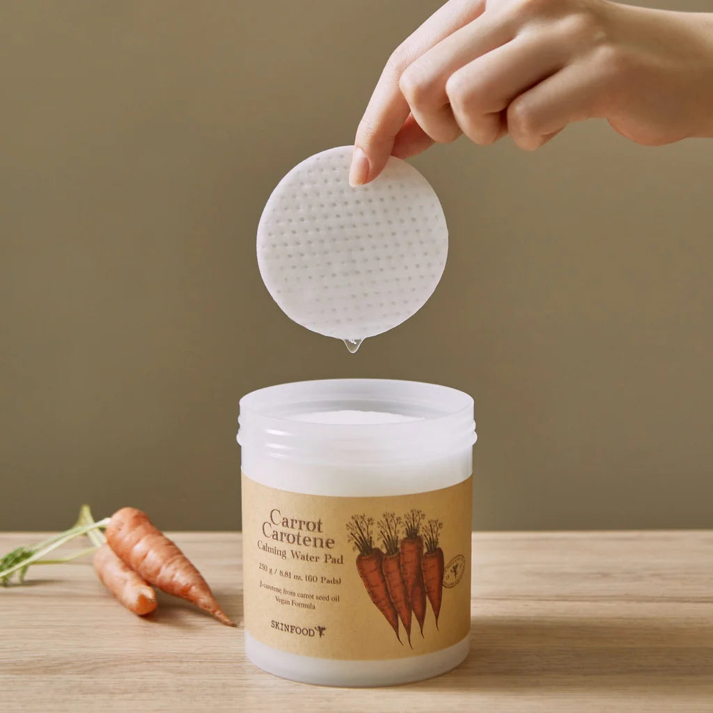 Disques apaisants Carrot Carotene Calming Water Pads SKINFOOD, main sortant un disque du pot, carottes à côté