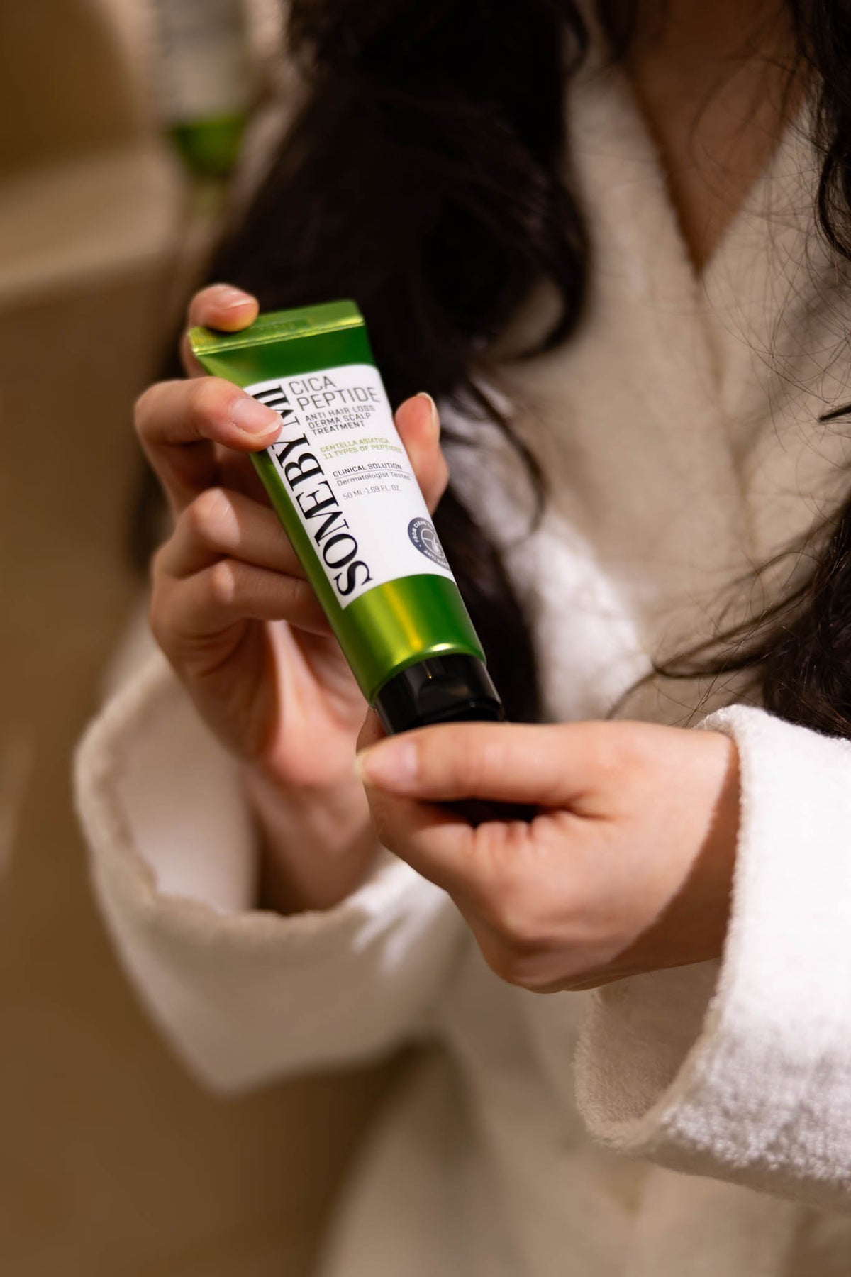 Soin cuir chevelu Cica Peptide Anti Hair Loss Derma SOME BY MI, tube vert tenu à deux mains par une mannequin en peignoir