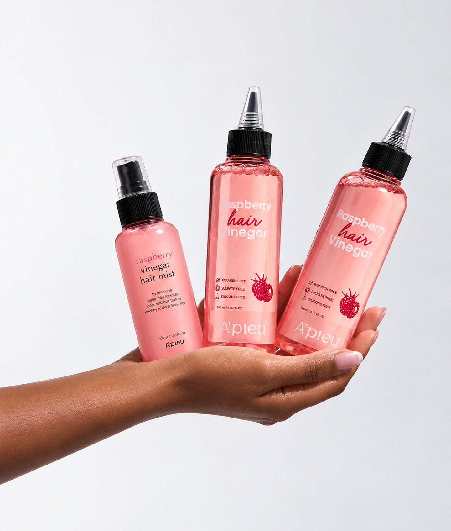 A’PIEU, une main présente trois flacons roses Raspberry Hair Vinegar sur fond gris clair