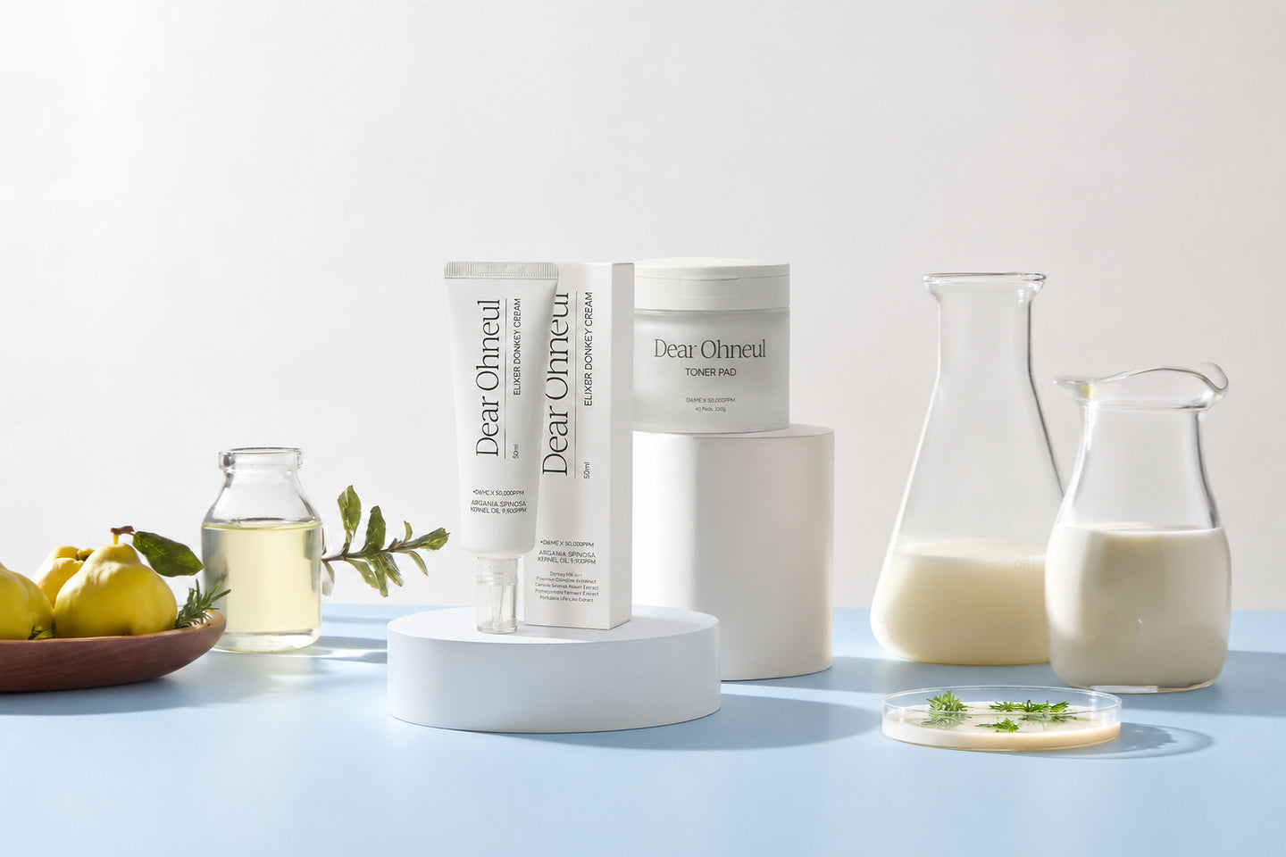 Dear Ohneul, tubes et pot de la marque sur des socles blancs, entre fruits jaunes et carafes de lait