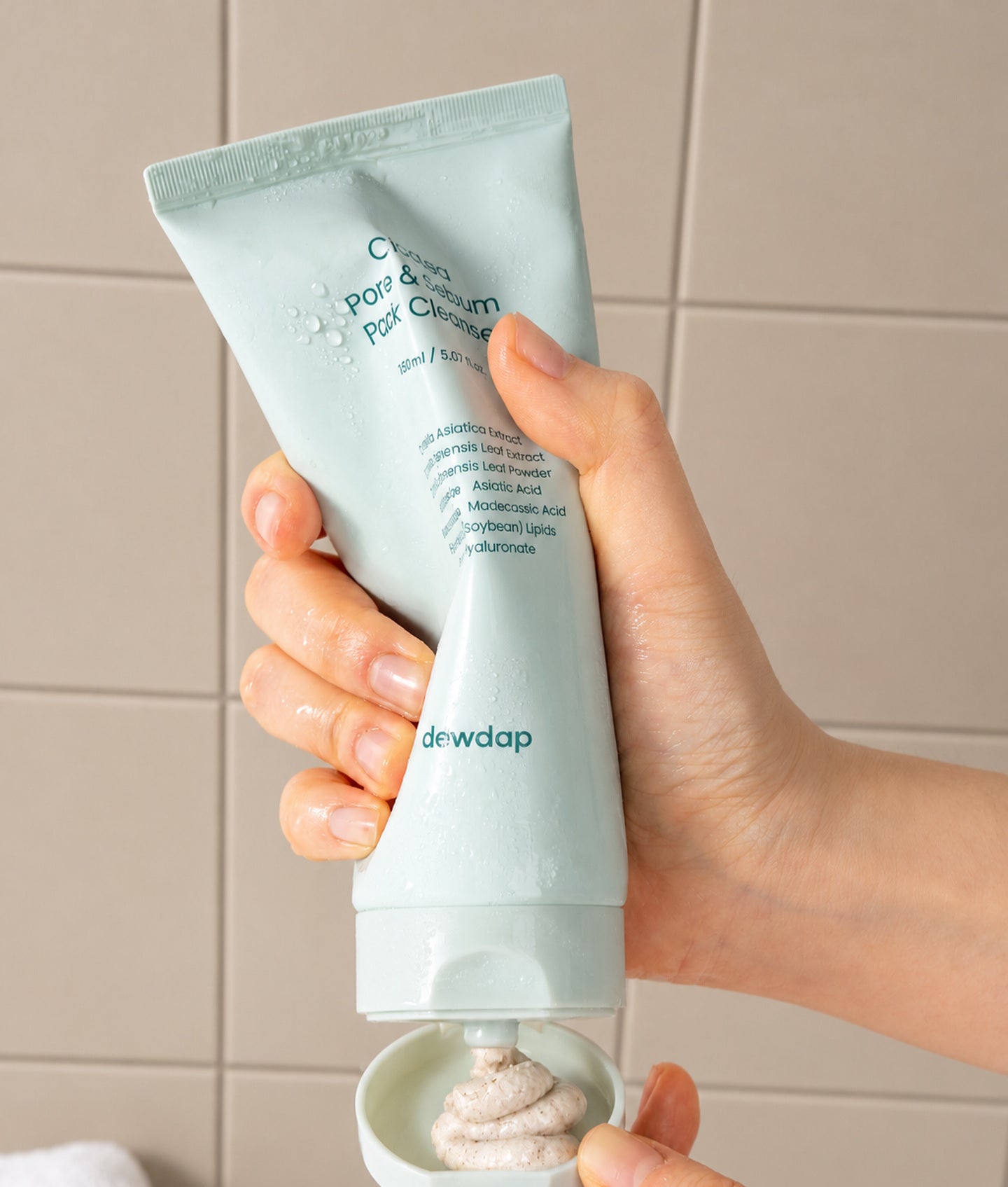 DEWDAP, une main presse un tube vert d’eau de Pack Cleanser devant un mur carrelé