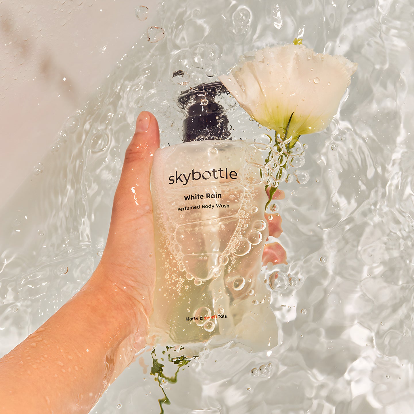 SkyBottle, une main tient un flacon White Rain Perfumed Body Wash dans l’eau, près d’une fleur blanche